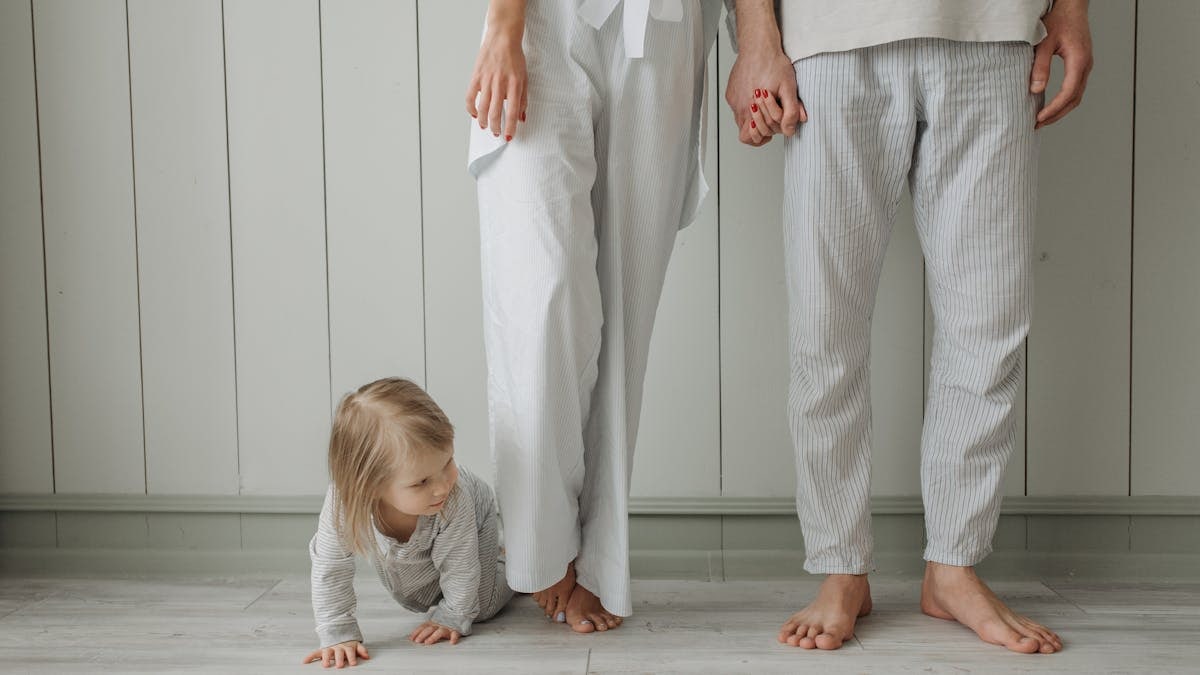 A toddler crawls on the floor while parents stand holding hands in a cozy home setting wearing casual clothes.